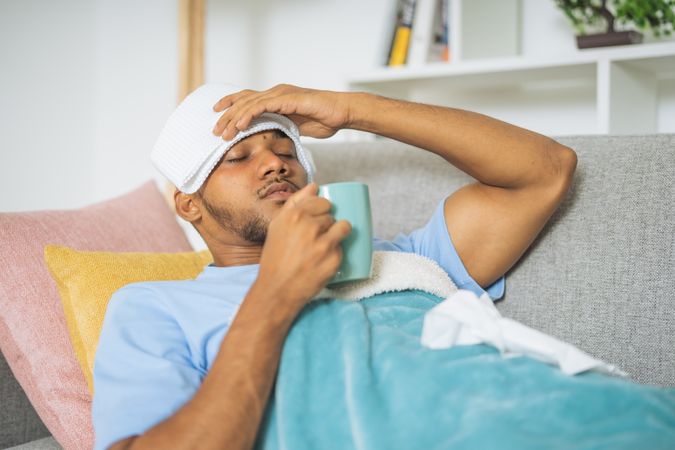Young man with fever, drinking a cup of tea, lying down on the couch