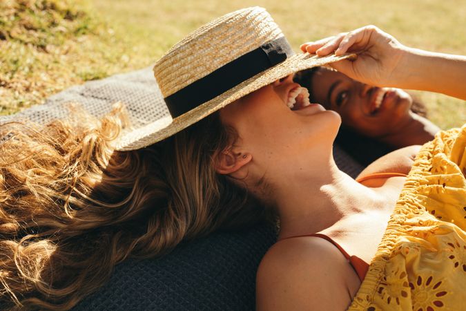 Close up of a cheerful woman covering her face with a hat relaxing outdoors in a park with friend