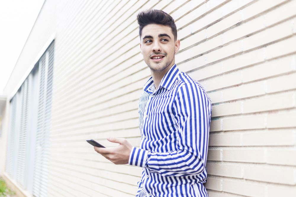 Side view of man leaning on house wall while checking his smartphone