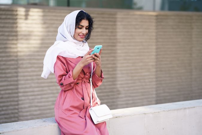 Female in headscarf and pink trench coat sitting outside with her phone