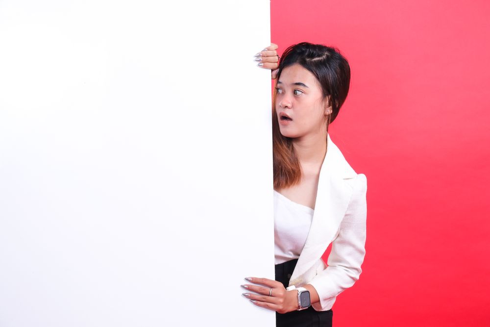 A woman wearing a jacket and a skirt is holding a large cardboard while looking to the side, isolated on a red background