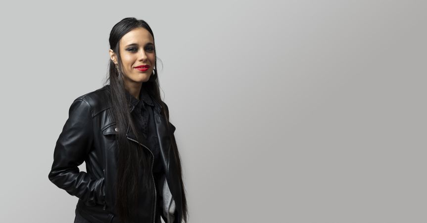 Portrait of a gothic rocker-looking woman, looking directly at the camera, wearing a leather jacket, on plain gray background