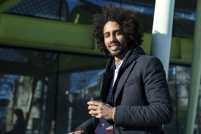 Portrait of cheerful handsome Black man in casual wear posing for the camera with take away coffee outdoors