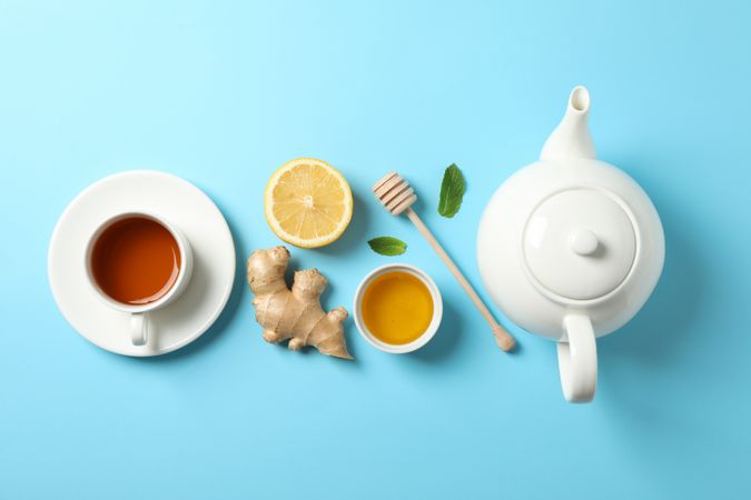 Flat lay. Cup of tea, teapot, lemon, ginger, mint, honey and dipper on blue background, copy space