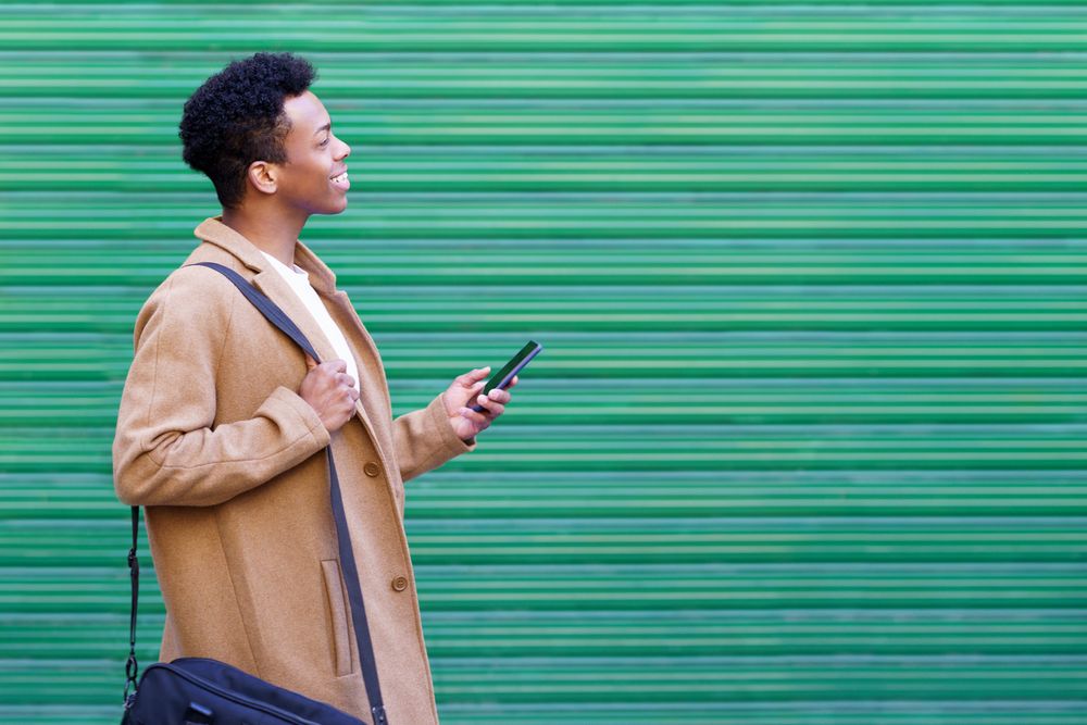 Side view of happy Cuban male texting on his phone in front of green wall
