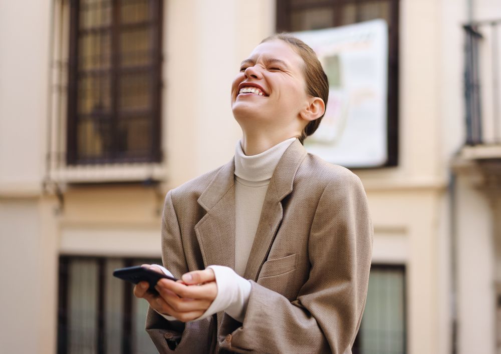 A Joyful and Young Woman Laughing While Enjoying Using Her Smartphone Outdoors in the Sun