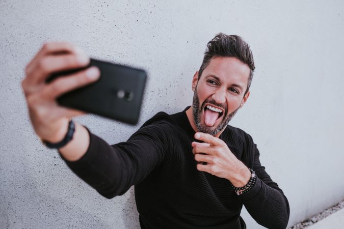 Adult stylish smiling man taking selfie on smartphone near grey wall