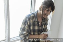 Female in flannel shirt writing in notebook leaning on window 5pXvx5