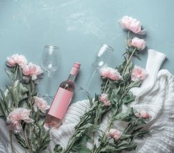 Bottle of rose wine with wineglasses on blue background with knitted sweater and many pink pale peonies flowers. Top view. Beauty
