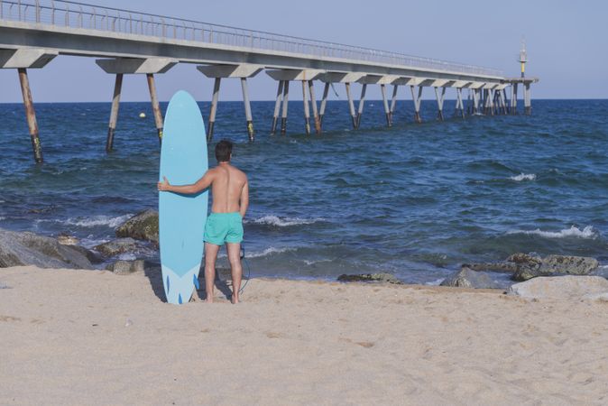 Full body shot of male surfer’s back standing in the rocks on the coast near pier