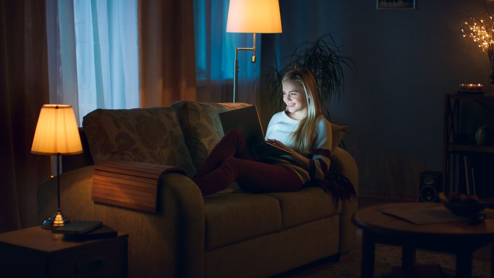 In the Evening Young Woman Lies on the Couch with a Laptop on Her Knees In Cosy Room