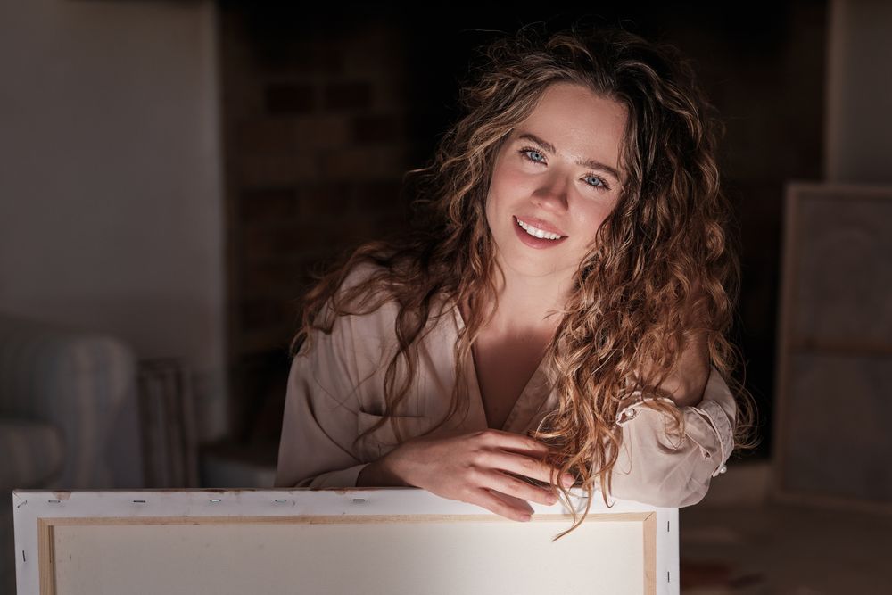 Smiling woman with long curly hair sitting near canvas