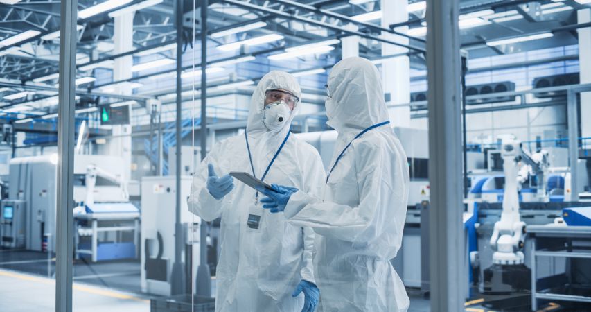 Industrial Scientists Wearing Disposable Protective Suits, Masks, and Goggles at a Production Plant. Specialist Using Tablet Computer, Analyzing Potential Work Hazards and Contamination at a Workplace