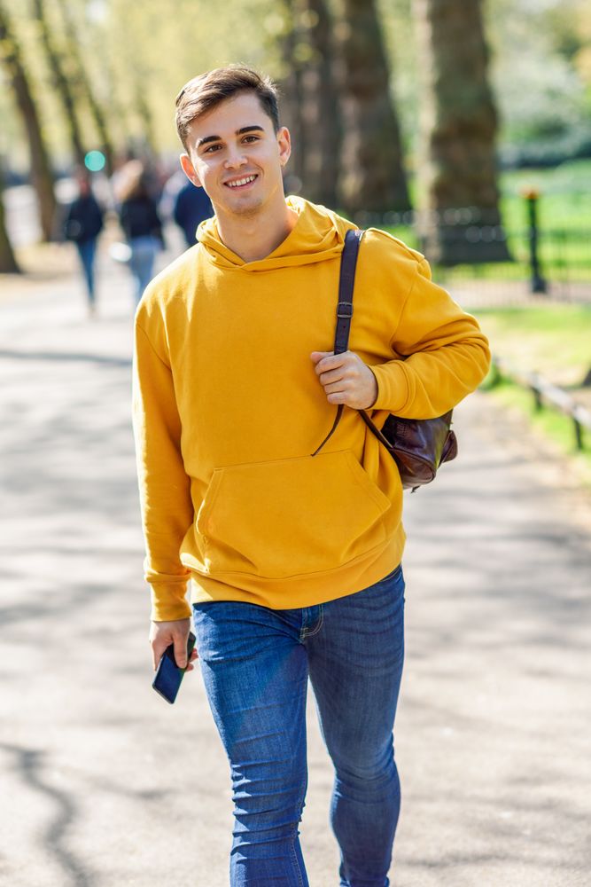 Smiling man in yellow hoodie with backpack walking in park, vertical