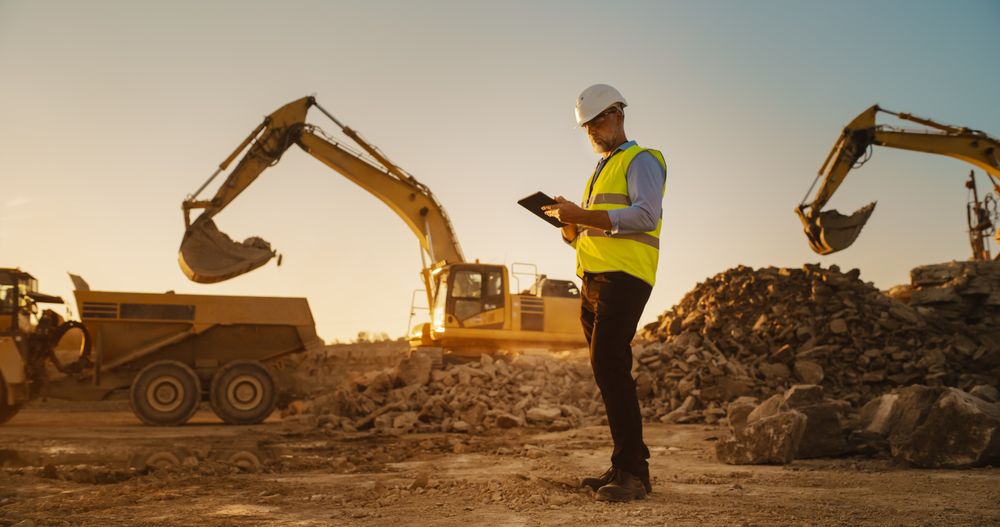 Male Real Estate Investor Wearing Protective Goggles And Using Tablet On Construction Site On a Sunny Day. Man Inspecting Building Progress. Excavator Loading Materials Into Truck.