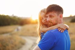 Man carrying daughter in long grass field at dusk