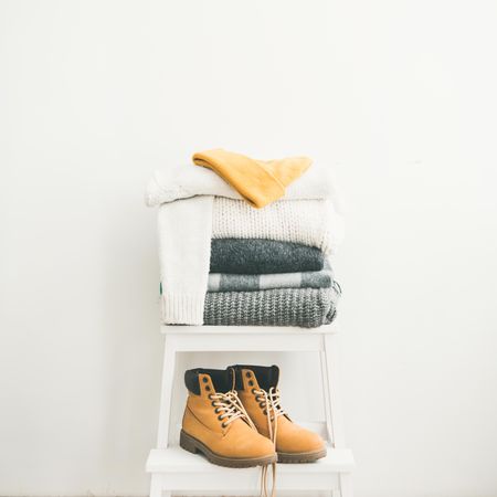 Pile of clean, folded sweaters on light background with yellow boots, square crop, copy space