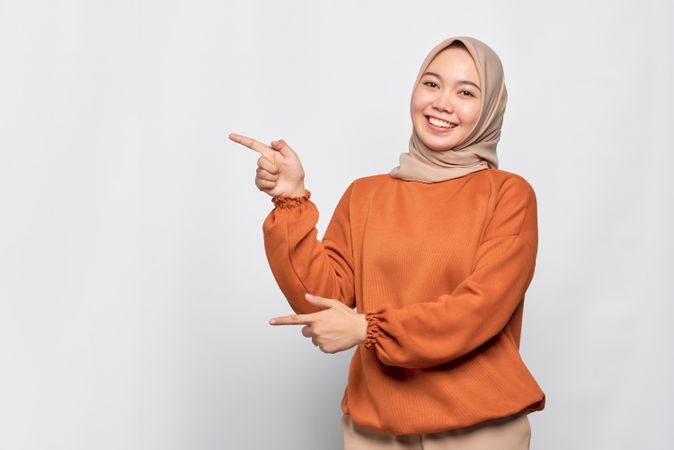 Merry Muslim woman in headscarf and orange sweater pointing to her side with both hands