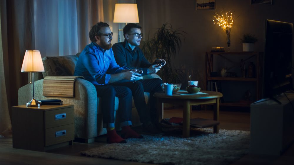 In the Evening Two Friends are Sitting on a Sofa in the Living Room and  Playing Competitive Video Games.