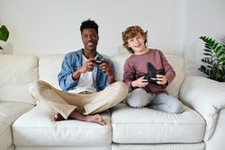 Cheerful Black man and young white boy with gamepads resting on sofa