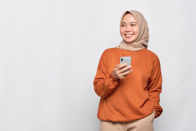 Calm Muslim woman in headscarf and orange shirt holding phone and looking at copy space