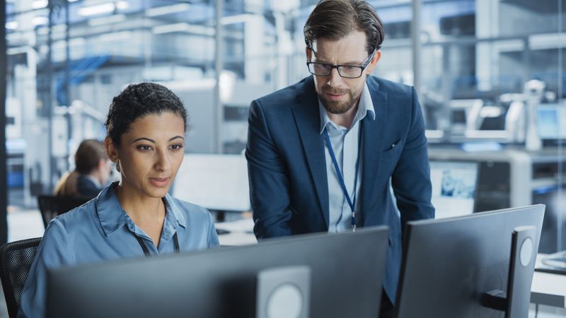 Portrait of a Young Male Team Leader and Hispanic Female Engineer Collaborating on a Project, Using Computer, Talking in a Factory with Robotic Arms Producing Devices for Technological Industries.