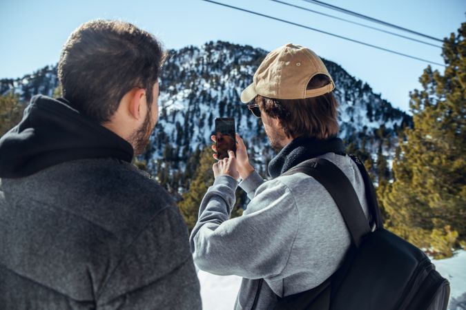 Men with smartphone and camera on hill between trees in winter