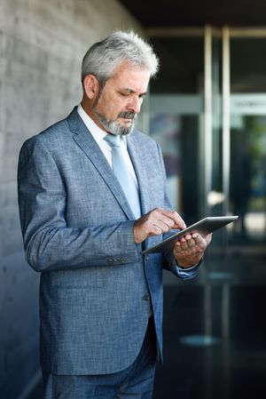 Grey haired man in formal suit checking tablet next to grey brick wall