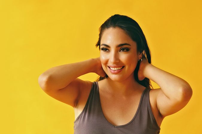 Headshot of Hispanic woman looking in distance with both hands at back of head on yellow background