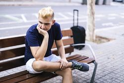 Young Man sitting on a bench with suitcase in the street with his hands on his face worried