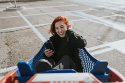 Smiling woman with smartphone in shopping trolley in parking lot