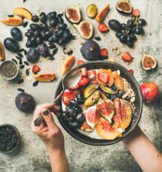 Woman with spoon in yogurt bowl over table of figs, grapes, peaches, and chia seeds, square crop