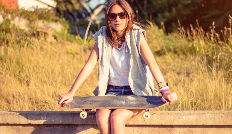 Young girl with skateboard sitting over the wall on summer