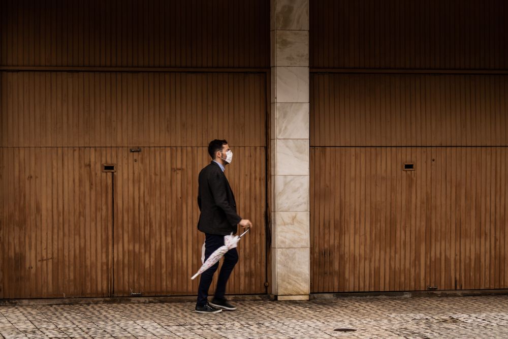 Side view of a man in suit with facemask holding an umbrella walking on sidewalk