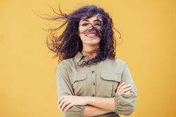 Female in army green jumpsuit smiling in front of mustard wall with arms folded and hair in face