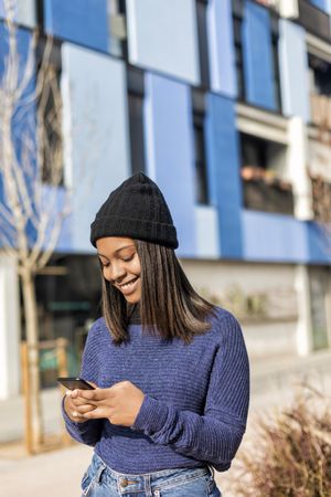 Female in hat and sweater checking phone outside blue building, vertical