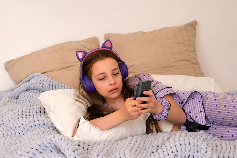Girl lying in bed with headphones, using smartphone.