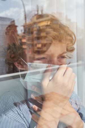 Boy covering face with medical mask near window