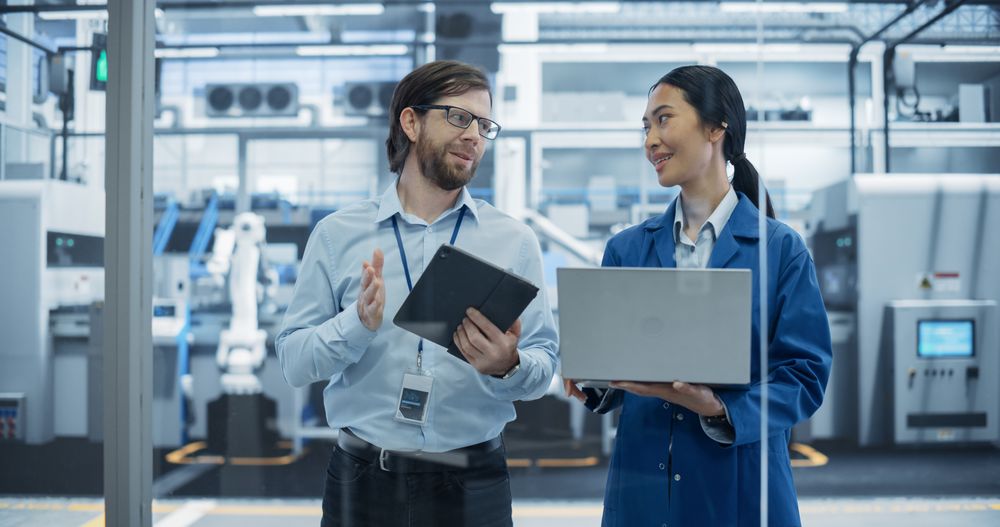 Engineer and Asian Female Project Manager Standing in Office, Talking and Using Computers and Monitoring Production at a Modern Automated Electronics Manufacture with AI Robotic Arms