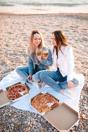Two mature women eating pizzas and drinking wine sitting on sand beach