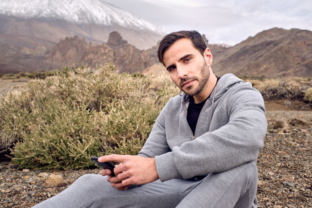 Young man sitting on grassy rocky ground and browsing smartphone