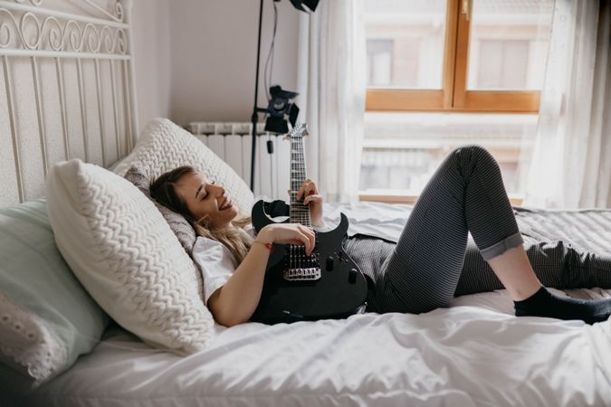 Side view of young brunette woman playing guitar on bed