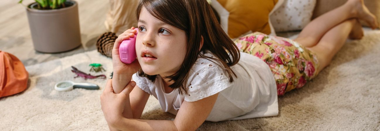 Banner of little girl talking with toy phone lying over rug surrounded by cushions at home