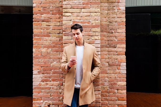 Front view of male in camel coat texting on phone while leaning on brick wall
