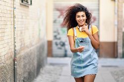 Excited Arab woman with headphones and curly hairstyle walking outdoors in lane with graffiti