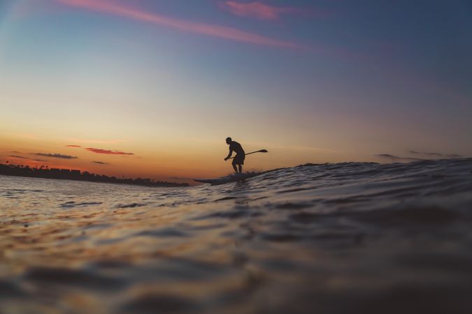 Man with paddle on surf board between waving water at sunset