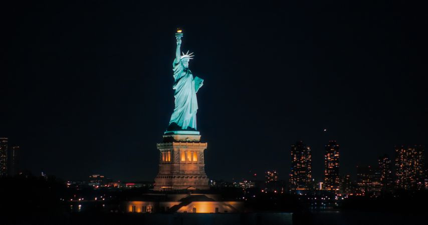 Aerial View of the Illuminated Statue of Liberty. Iconic Travel Destination and Tourist Attraction with Manhattan and Brooklyn Office Buildings All Lit Up at Night. Nightlife in New York City