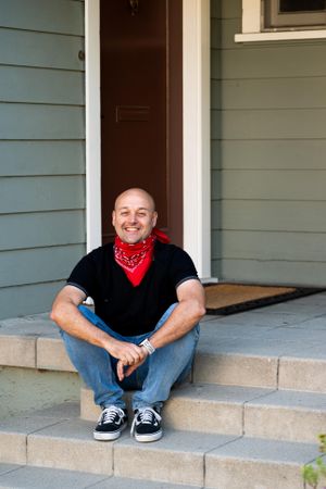 Man with red bandana around his neck sitting on steps in front of house smiling