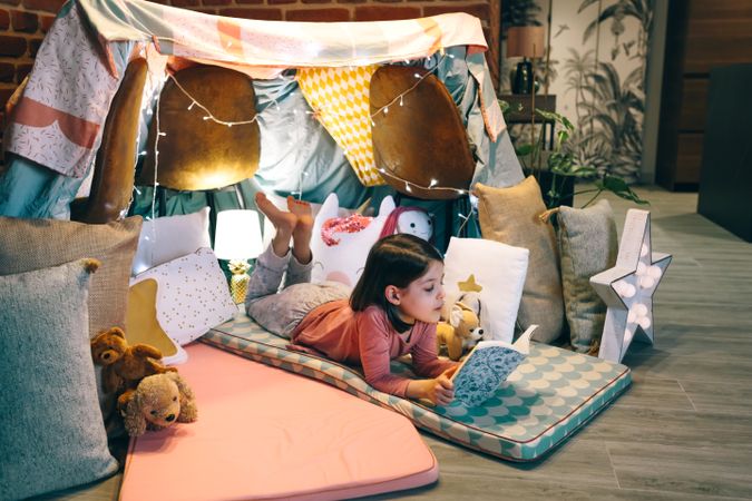 Little girl reading and relaxing lying over sleeping mat in a cozy tent at home