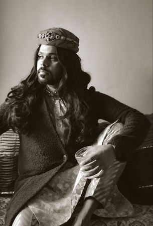 Grayscale photo of Indian man with long hair sitting on floor holding a drink